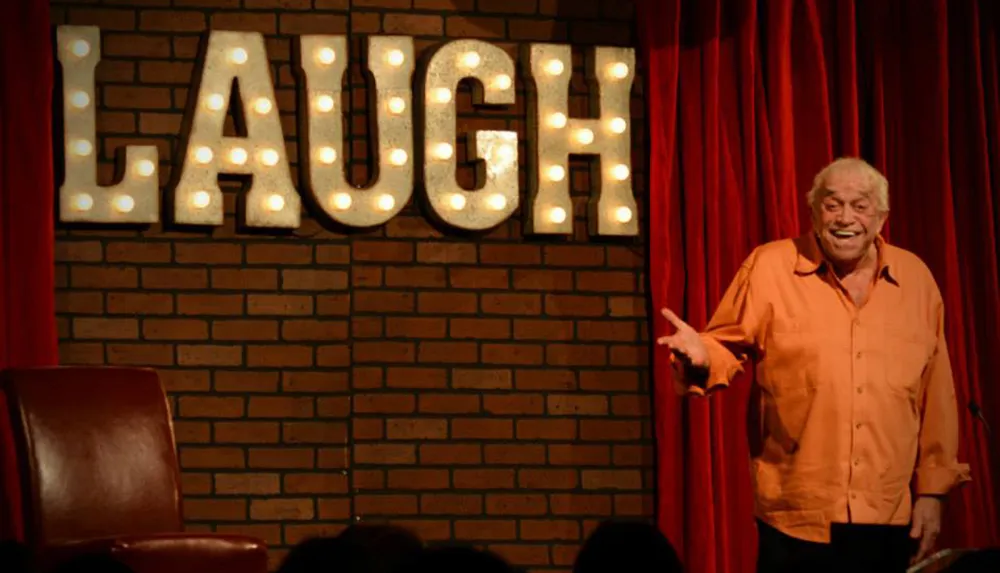 A smiling person is performing on stage at a comedy club with a lit-up sign that reads LAUGH in the background