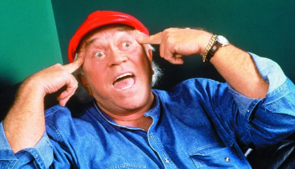 A man in a red cap and a denim shirt is making a surprised or goofy face while pulling his fingers near his temples