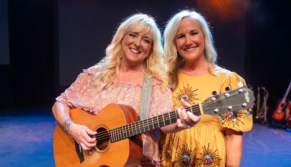 Two smiling women are standing on a stage one holding a guitar appearing to be musicians enjoying a performance moment