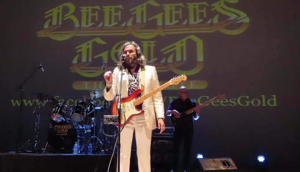 A person holding a guitar stands on stage with the Bee Gees logo projected in the background accompanied by another musician and musical equipment