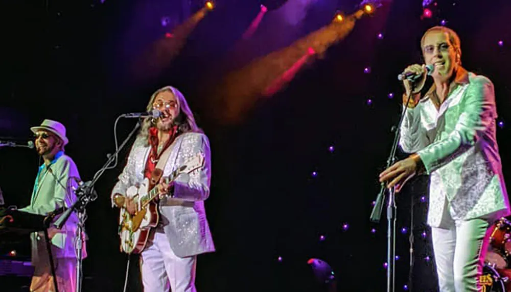 Three musicians perform on stage with colorful lighting in the background with one singing into a microphone another playing a guitar and the third on keyboards