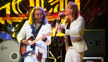 A male guitarist and a female singer perform on stage against a vibrant, multicolored backdrop, with the guitarist sporting a white jacket and the singer in a matching ensemble.