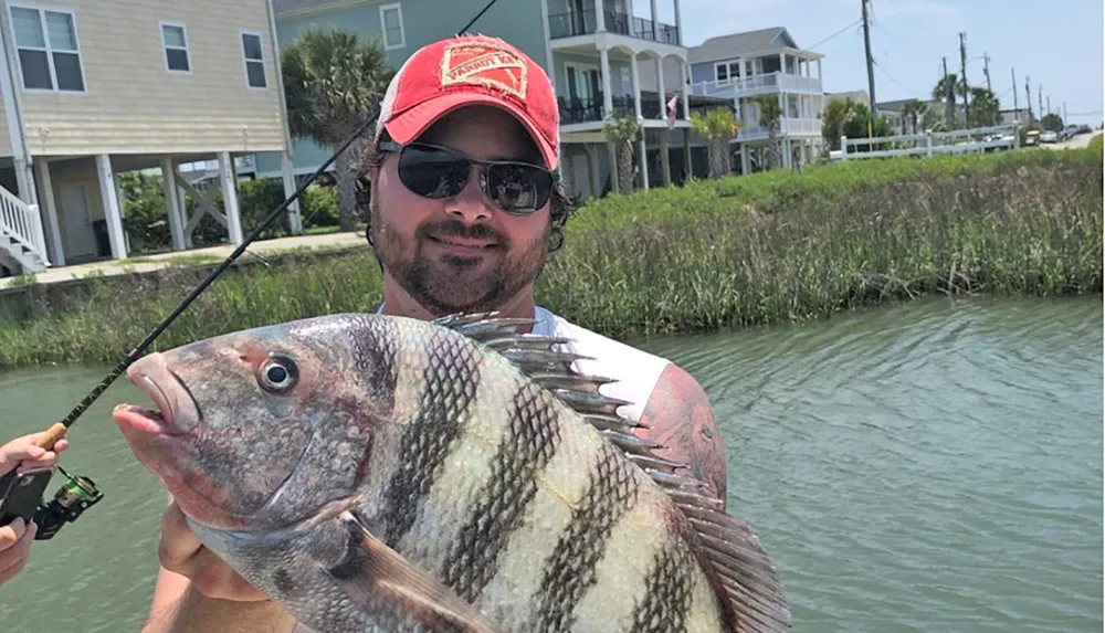 A man wearing sunglasses and a cap smiles as he holds up a large fish with a fishing rod in hand by a waterway with houses in the background