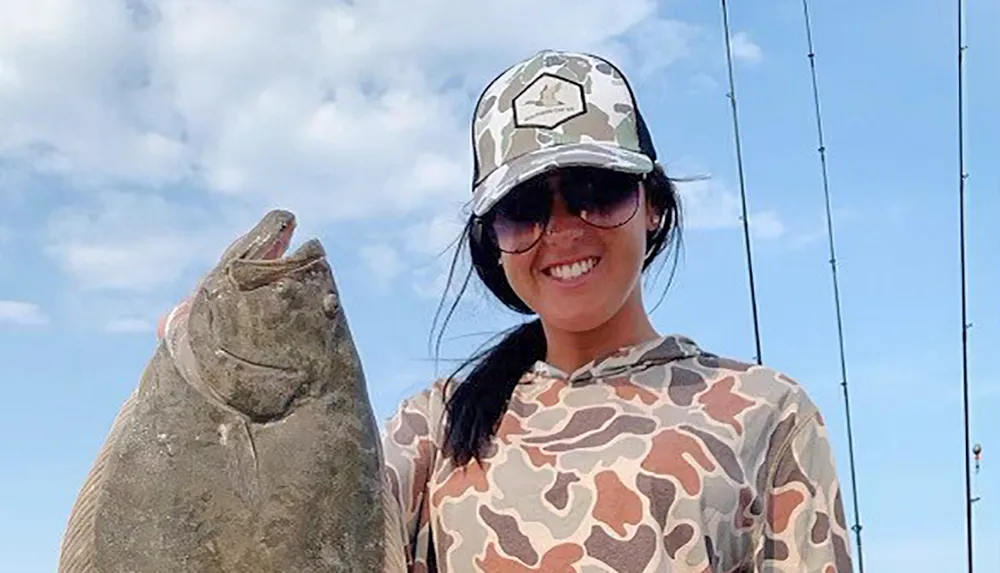 A smiling person wearing a camouflage pattern hat and shirt is holding a large fish with fishing rods visible in the background