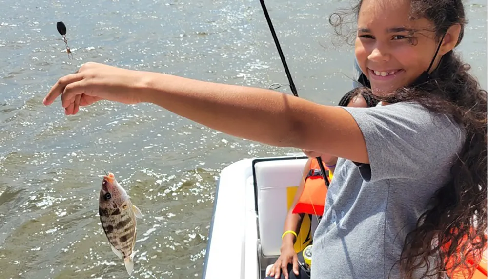 A smiling girl is holding a fishing rod with a small fish at the end of the line while on a boat