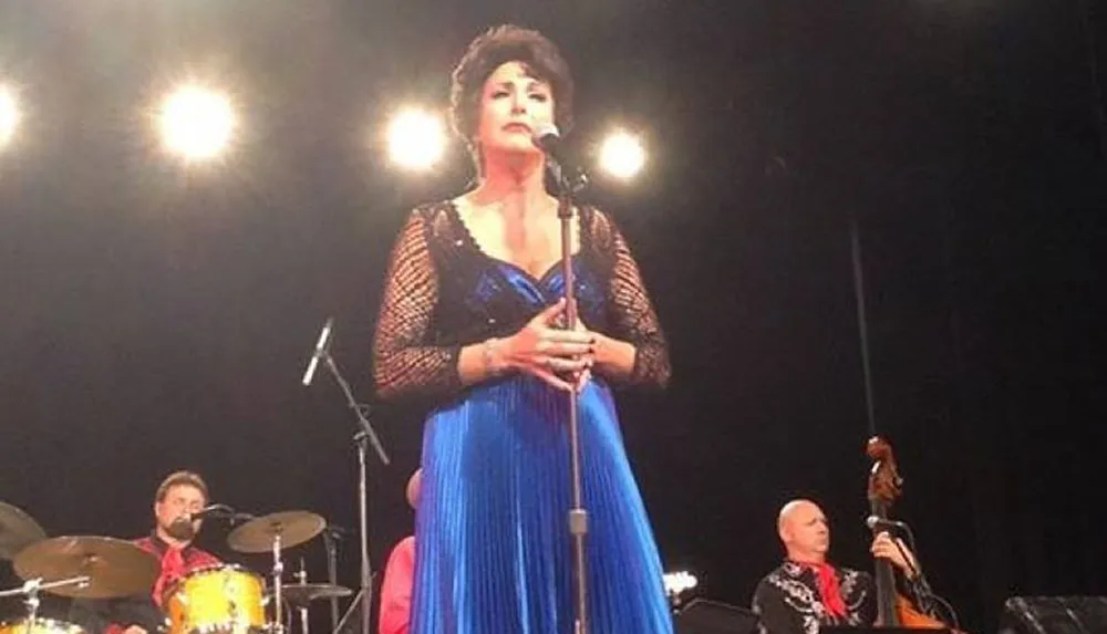 A performer in a blue dress stands on stage holding a microphone with musicians including a drummer and a bassist in the background