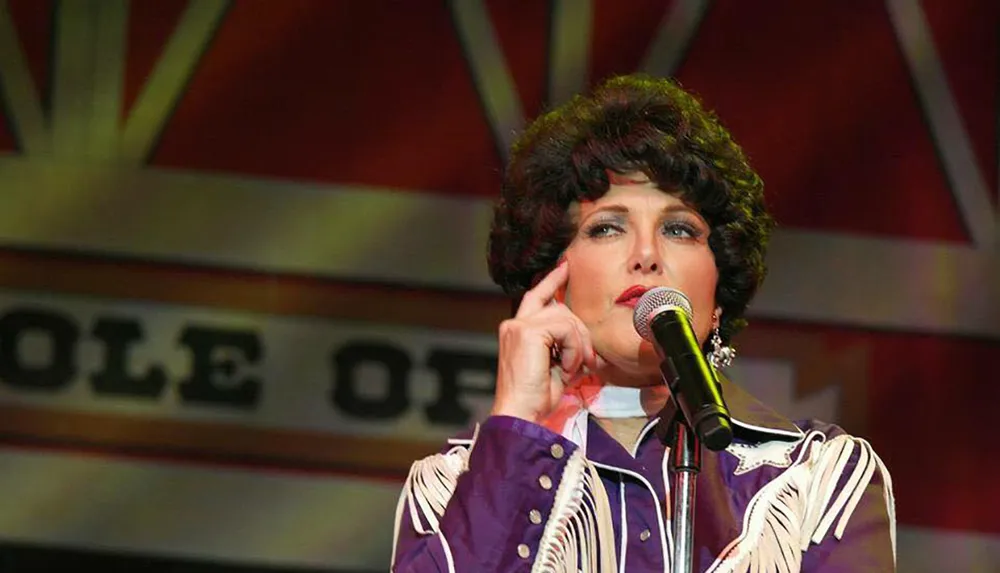 A performer wearing a Western-inspired outfit speaks into a microphone on stage with decor that suggests an old-fashioned music theme