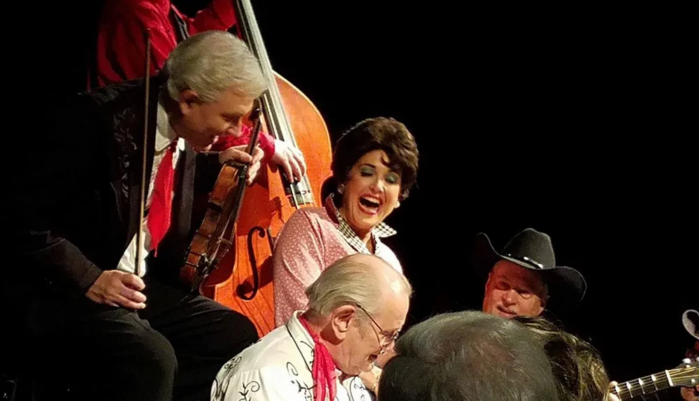 A group of performers including a laughing woman in a red checkered outfit a man playing upright bass and individuals wearing western-style attire are onstage likely engaging in a country or folk music performance