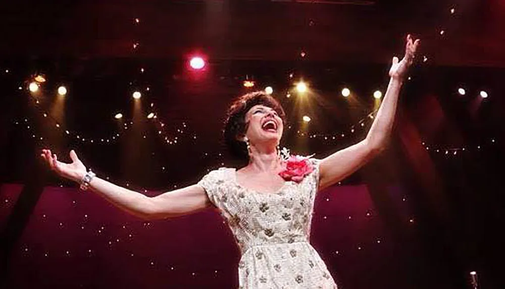 A woman in a sparkling dress with arms wide open is joyfully singing on a stage with lights blurred in the background