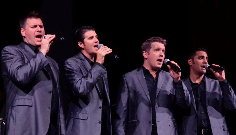 Four men in matching gray suits are singing passionately into handheld microphones on stage