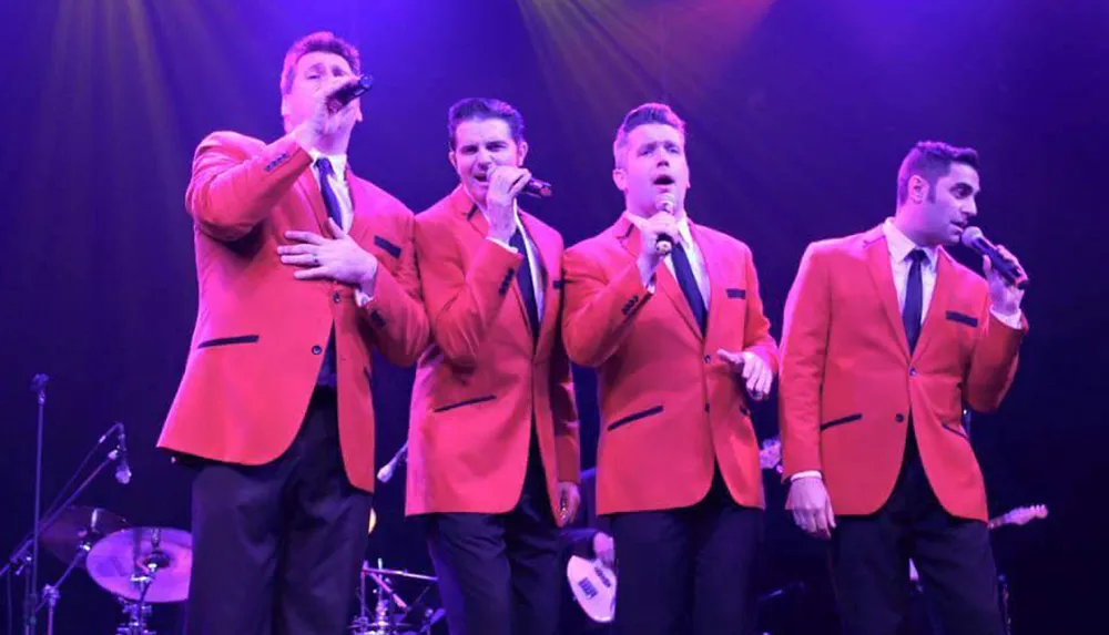 Four men in red suits are performing on a stage with microphones illuminated by stage lighting in the background