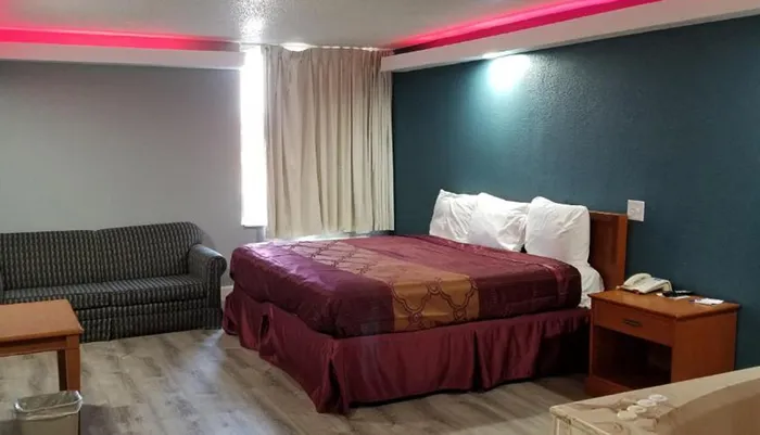 The image shows a simply furnished hotel room with a large bed a patterned sofa a small study desk and contrasting red ambient lighting along the ceiling