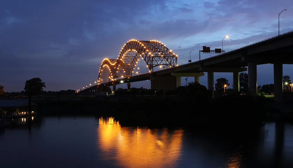 The image captures a lit-up arched bridge over a body of water at dusk with its reflection visible on the water surface and a twilight sky in the background