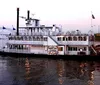 A traditional paddlewheel riverboat named Memphis Queen is navigating a river with a bridge and a pyramid-shaped structure in the background