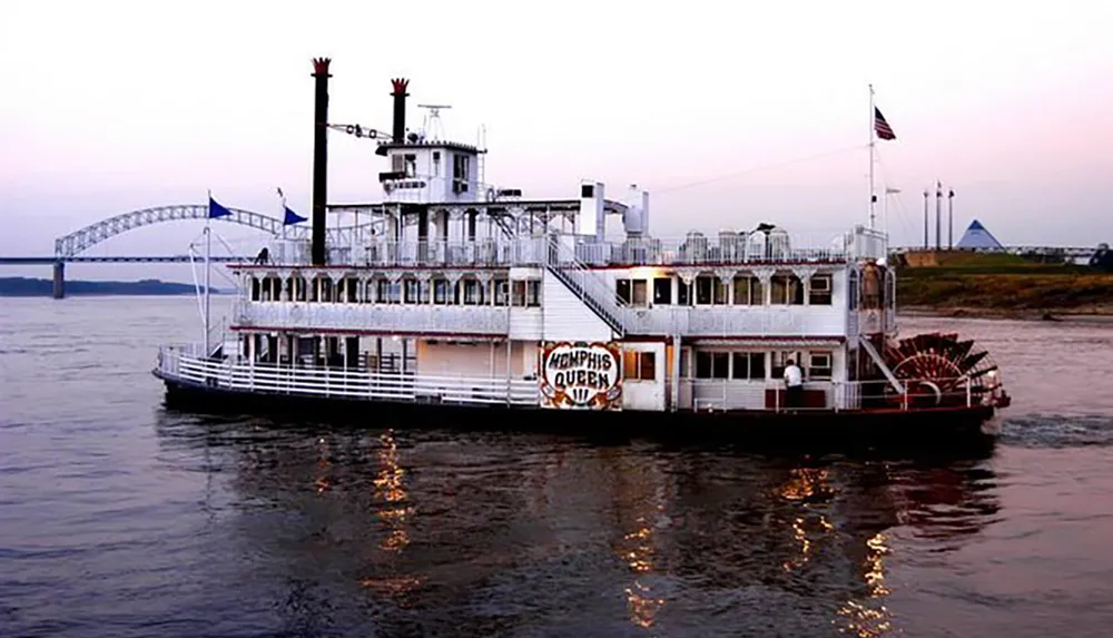 A traditional paddlewheel riverboat named Memphis Queen is navigating a river with a bridge and a pyramid-shaped structure in the background