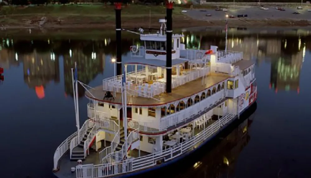 A paddle steamer with multiple levels is docked on a calm river at dusk with its lights reflecting off the waters surface