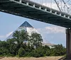 A traditional paddlewheel riverboat named Memphis Queen is navigating a river with a bridge and a pyramid-shaped structure in the background