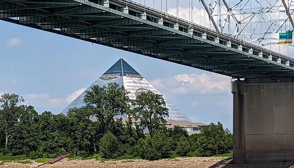 A large glass pyramid structure is visible beneath a green metal bridge with trees and a clear sky in the background