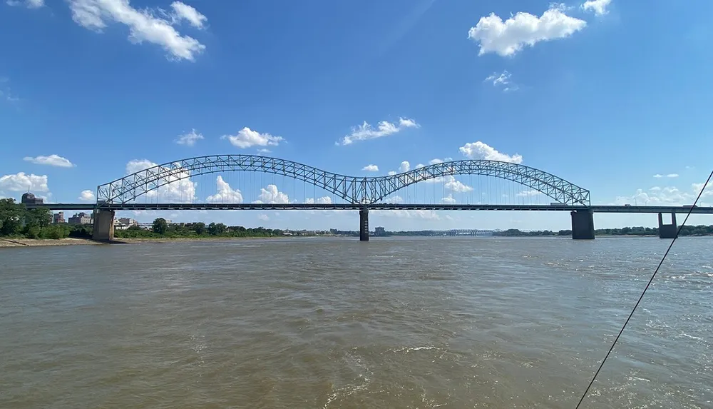 The image features a large steel arch bridge spanning a wide muddy river under a partly cloudy sky