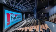 The image shows an empty modern theater with a large screen displaying 