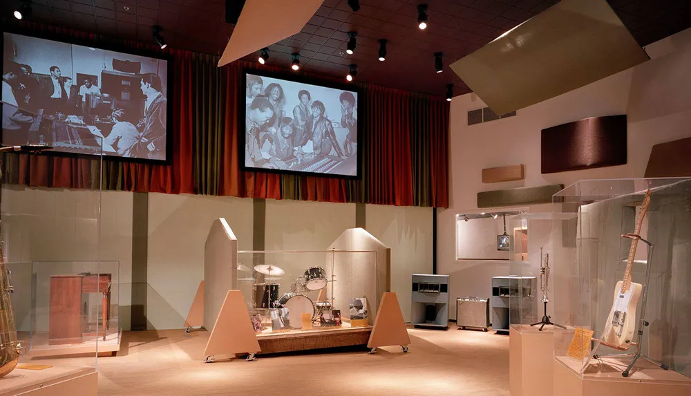 This image shows a music exhibition with historical instruments displayed in glass cases and large vintage photographs projected on screens providing a retrospective ambiance