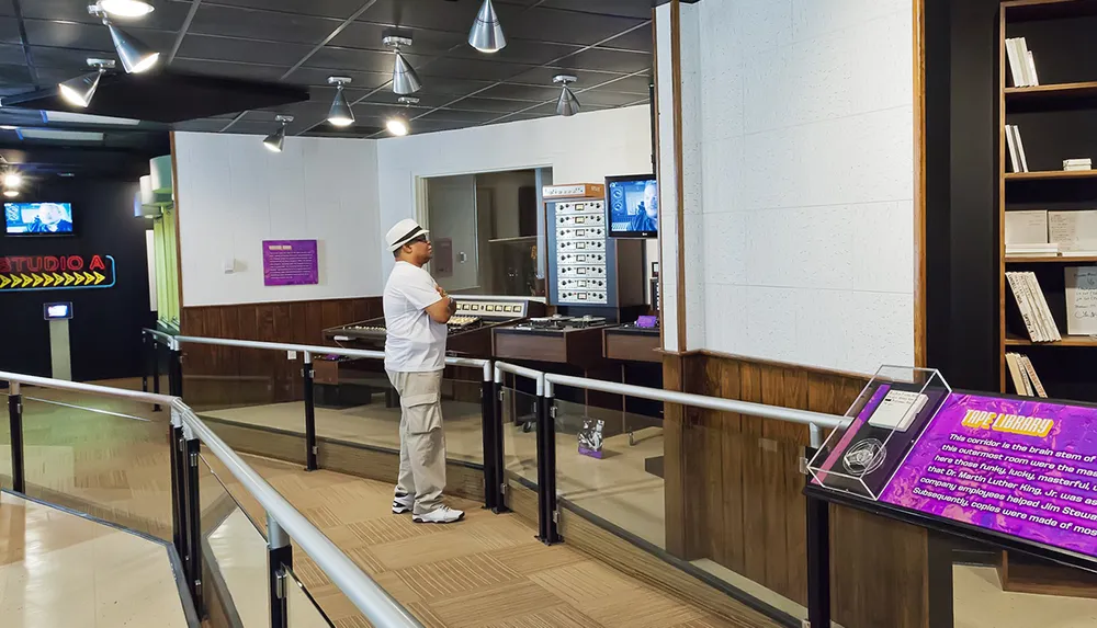 The image shows an individual observing a vintage mixing console in a music studio exhibit with informative signs visible like STUDIO A and TAPE LIBRARY