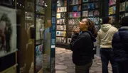 A person appears engrossed in looking at a collection of items on display in a museum exhibit, with others around her also viewing the displays.