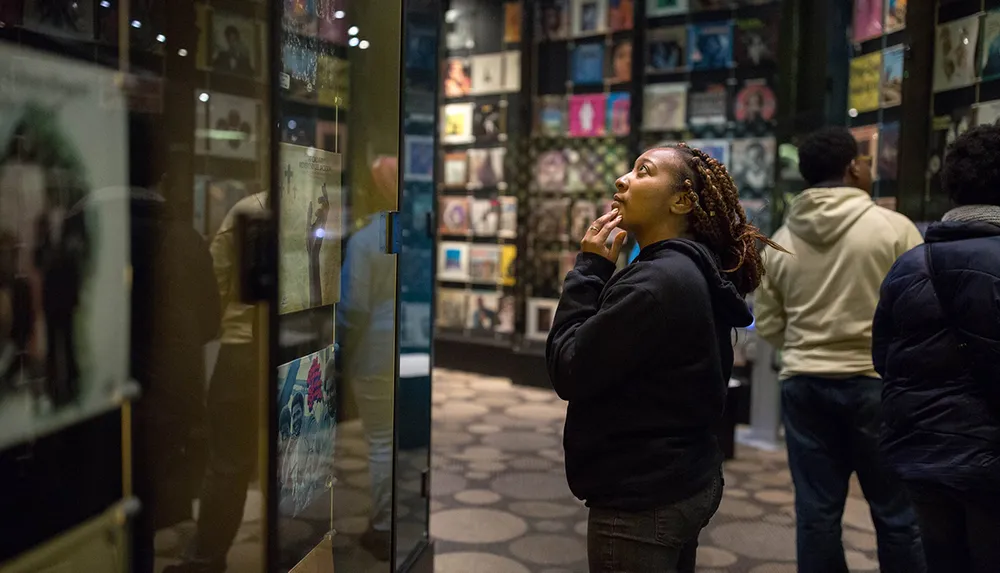 A person appears engrossed in looking at a collection of items on display in a museum exhibit with others around her also viewing the displays