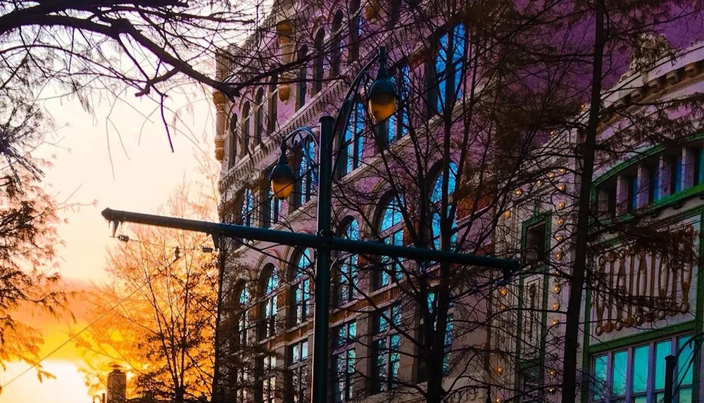 The image shows a vibrant urban street scene at dusk with the silhouette of a street lamp and bare tree branches set against a colorful building and sunset