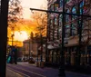 This is an image of a city street at dusk with the warm glow of the sunset visible between the silhouettes of trees and buildings creating an atmospheric urban scene