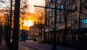 This is an image of a city street at dusk, with the warm glow of the sunset visible between the silhouettes of trees and buildings, creating an atmospheric urban scene.