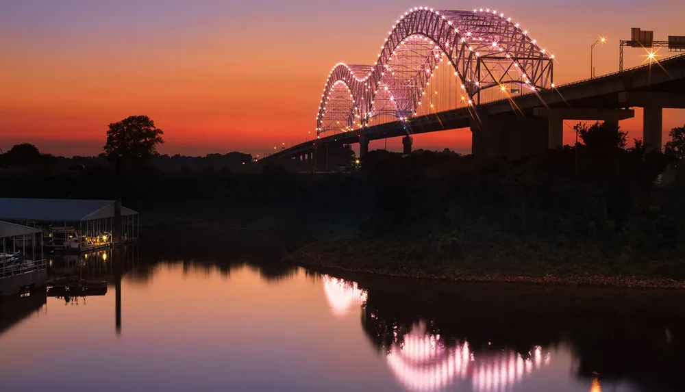 A lit-up arched bridge spanning a calm river reflects in the water below during a colorful sunset