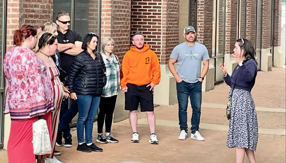 A group of people is listening to a woman who appears to be giving a talk or a tour outside a brick building