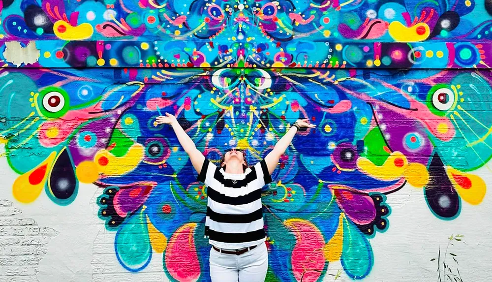 A person with arms raised stands in front of a vibrant colorful mural with abstract patterns and eye motifs
