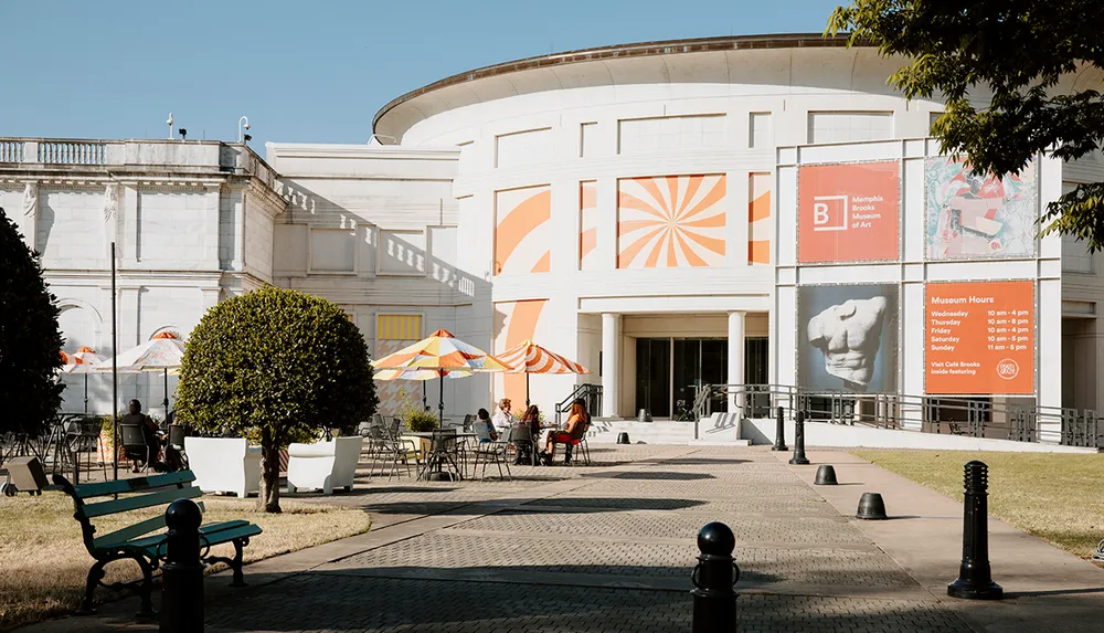 The image depicts the outdoor seating area of the Memphis Brooks Museum of Art with tables chairs and colorful umbrellas set against the museums architectural backdrop
