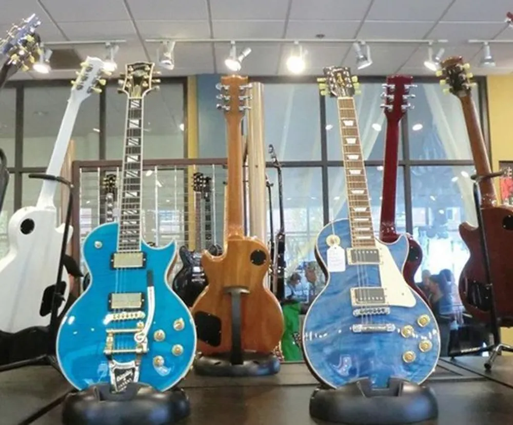 The image shows a collection of various electric guitars on display featuring different colors and designs with a reflective floor and large windows in the background of a music store