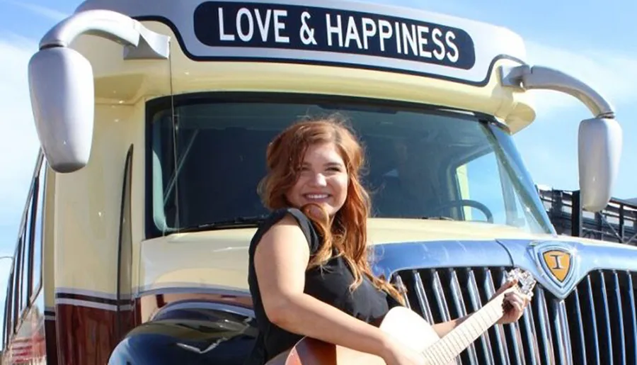 A smiling person holding a guitar is sitting in front of a cream-colored truck with the words LOVE & HAPPINESS on its sun visor.