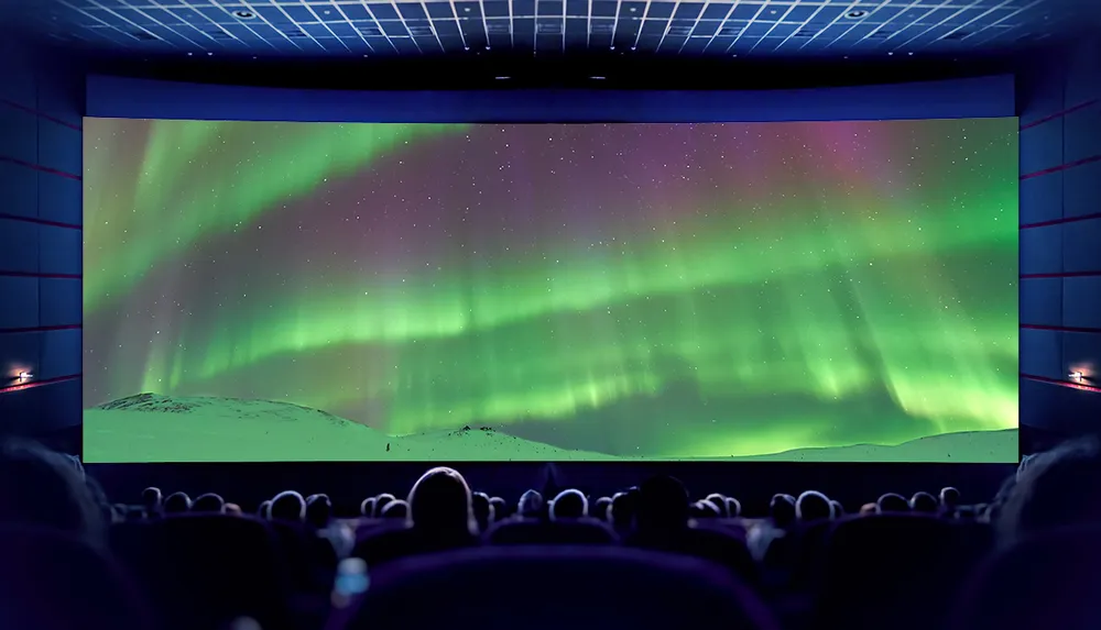 This image depicts an audience in a dark movie theater looking at a large screen displaying a vibrant scene of the northern lights above a snow-covered landscape