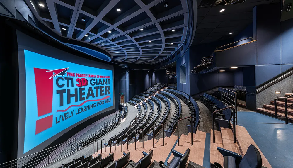 The image shows an empty modern theater with a large screen promoting the CTI 3D Giant Theater at the Pink Palace Family of Museums featuring dark blue walls comfortable tiered seating and a curvilinear overhead design