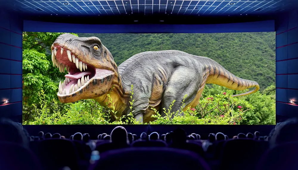 The image shows a movie theater from the perspective of the audience with a giant dinosaur displayed on the screen creating an immersive cinematic experience
