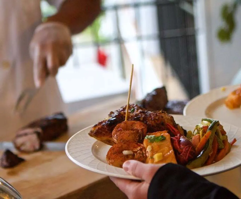 A person is holding a plate with grilled meat and vegetables with another person in the blurry background presumably slicing additional meat