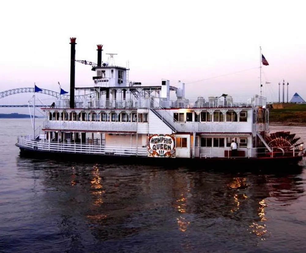 The image shows the Memphis Queen a classic Mississippi paddlewheel riverboat on the water at dusk with a bridge in the background