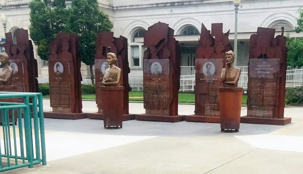 The image displays a series of brown sculptures resembling lecterns with busts and information plaques dedicated to historical figures set outdoors with a building and greenery in the background