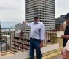 A man is pointing towards the horizon seemingly explaining something to a small group of people on a rooftop with a cityscape and a river in the background