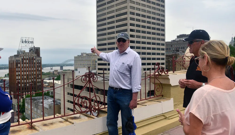 A man is pointing towards the horizon seemingly explaining something to a small group of people on a rooftop with a cityscape and a river in the background