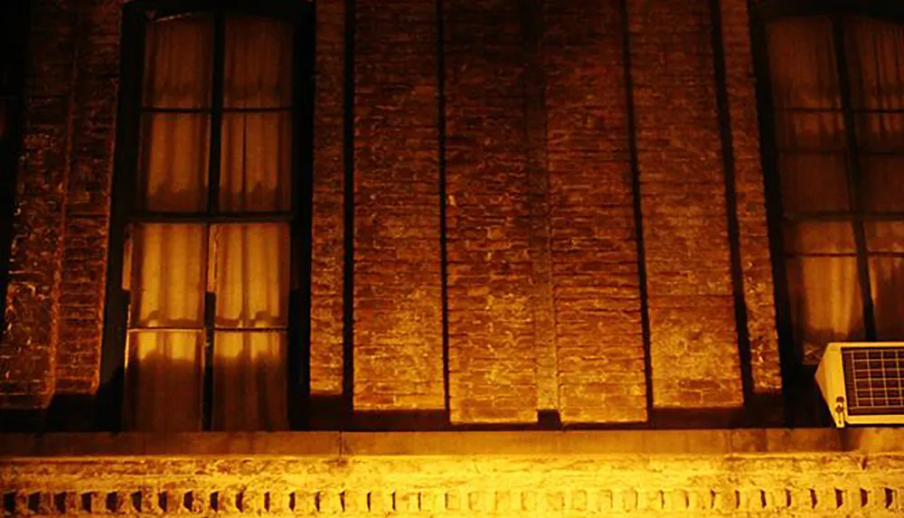 The image shows a warmly lit segment of a brick building at night featuring tall windows and an air conditioning unit on the right side