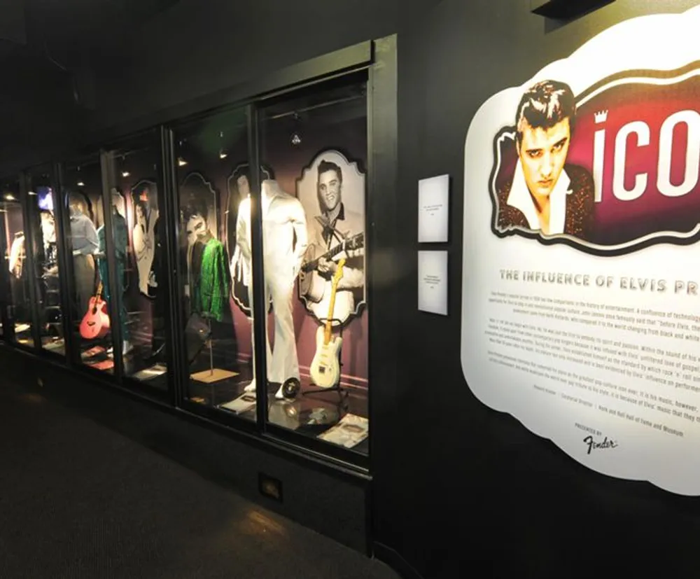 The image shows an exhibition featuring iconic outfits and guitars presumably influenced by or belonging to Elvis Presley as indicated by the graphic and text on the right