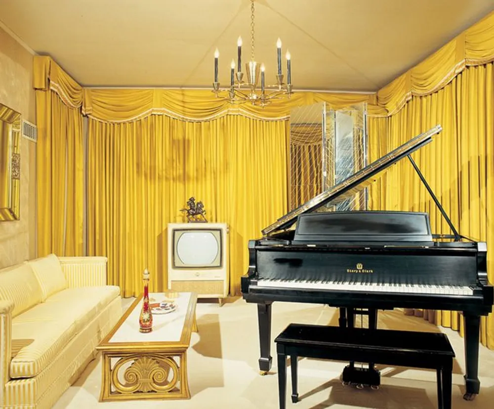The image features an opulent room dominated by yellow hues with grand curtains a chandelier a black grand piano a vintage television and elegant furniture