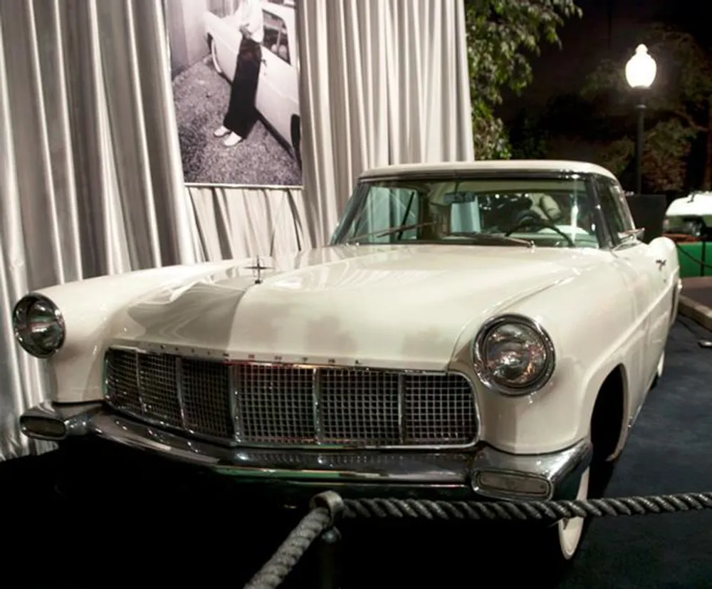 The image displays a classic white vintage car on display with a large black-and-white photograph in the background set against draped curtains with a roped-off area and a streetlamp-style light present