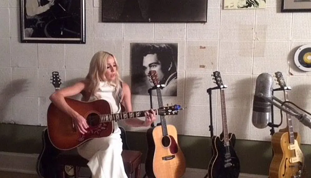 A woman is sitting and playing an acoustic guitar in a room adorned with musical instruments and a framed picture on the wall